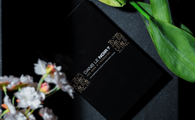 Dans le Noir ? black booklet surrounded by green leaves and white flowers on a dark stone background