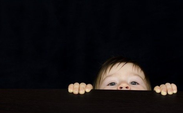 A child peeking curiously over a table in a dark setting, evoking a playful and reassuring family sensory experience in Hong Kong